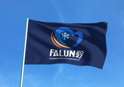 En flagga med logotypen för skid-vm 2027 i Falun syns mot en blå himmel.