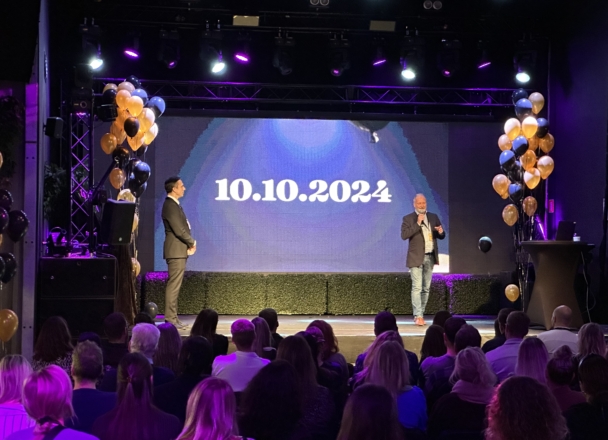 En fullsatt, nedsläckt lokal i Falun. På scenen står två personer och på skärmen visas datum för Panangs 10-årsfirande. Gula och svarta ballonger.